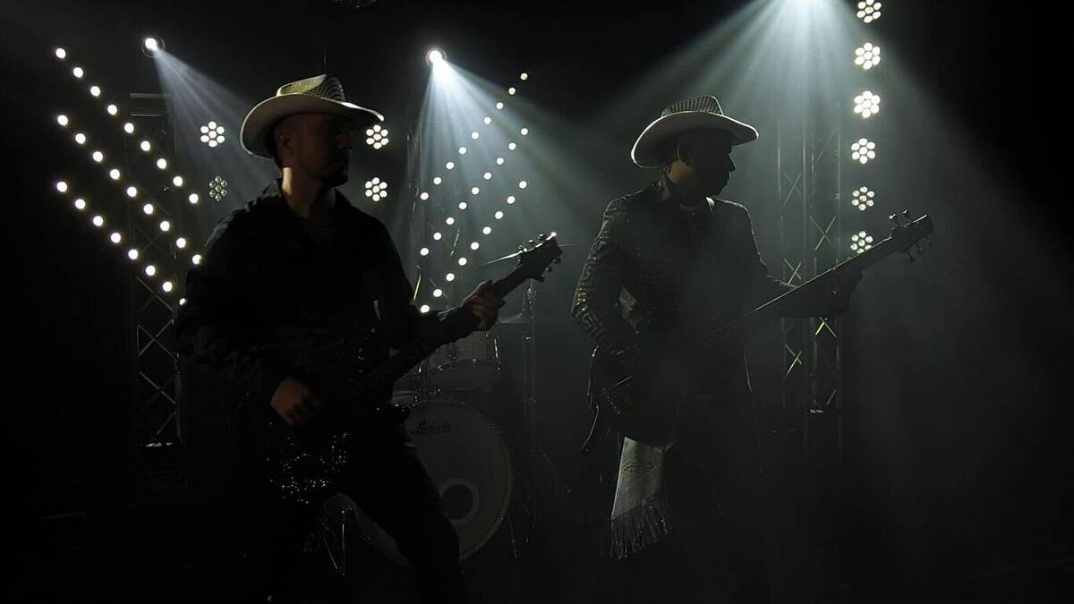 Two guitarists in cowboy hats performing in silhouette against dramatic white backlighting and matrix/blinder panels with heavy haze