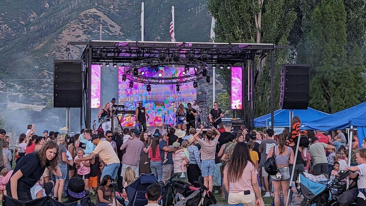 A busy daytime outdoor festival with a band on a purple/pink-lit stage, LED video panels, and a large dancing crowd against a mountain backdrop