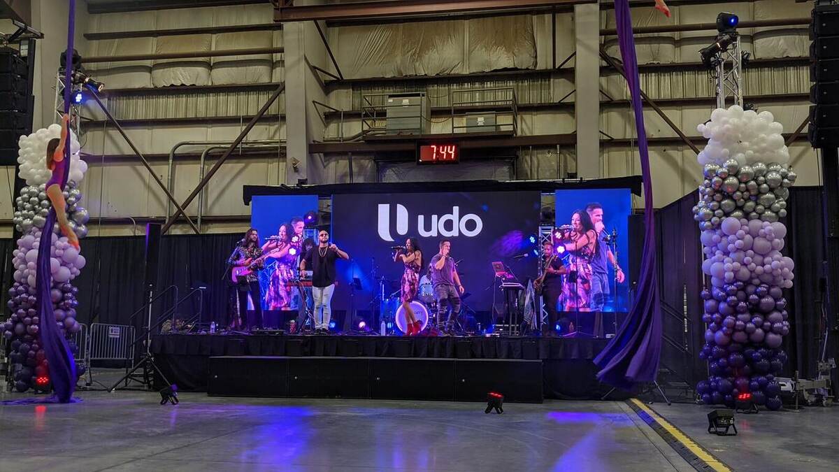 A live band performs on the 'udo' branded LED stage with purple lighting and balloon columns while a host addresses the crowd