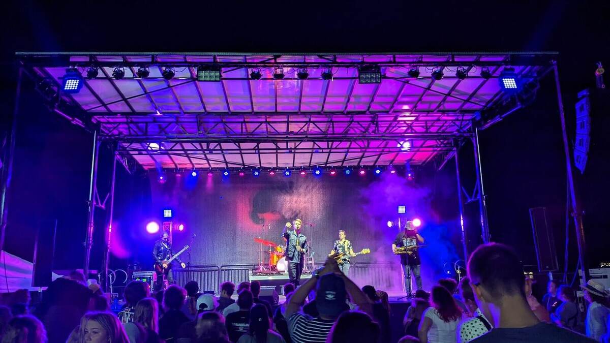 A band performs on an outdoor stage saturated in vivid purple and magenta wash lighting with haze, in front of a large night crowd holding up phones