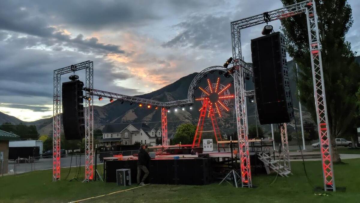Outdoor concert stage at dusk with truss goalpost rigging, line-array speakers, a circular lit backdrop element, and mountains behind under a dramatic sunset sky
