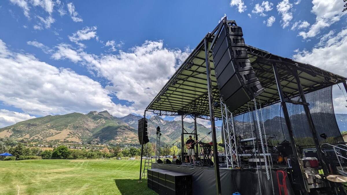 A large outdoor mobile stage with a line-array PA system and truss roof set up on a green field with dramatic mountains and blue sky behind it; a lone musician on stage during daytime