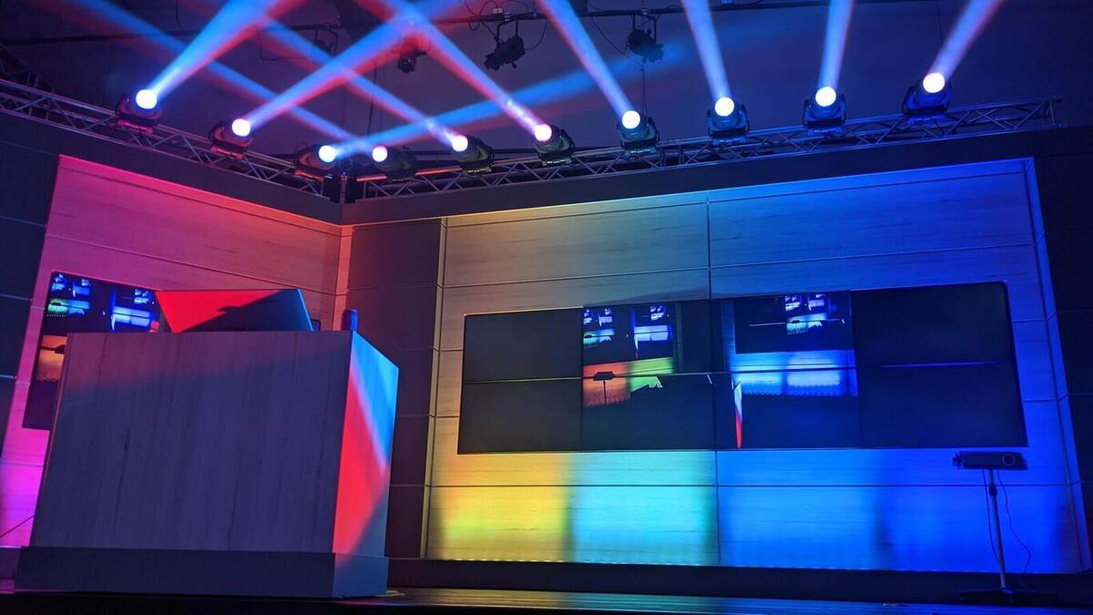 Corporate-style stage set with a wood-panel wall of mounted video monitors, a podium, moving-head beam lights overhead in blue and red, and colorful floor wash lighting